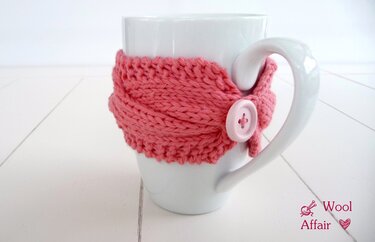 Weiße Tasse mit lachsrosa gestricktem Tassenwärmer mit Zopfmuster und Knopf