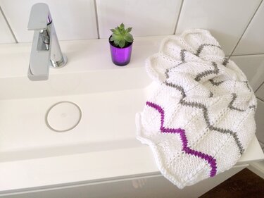 Weißes gestricktes Tuch mit grauen und violetten Zickzackstreifen auf dem Rand eines Waschbeckens neben kleinem Sukkulenten-Topf
