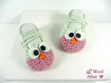 Zwei mintgrün-rosa gestrickte Babyschuhe mit Eulenaugen und orangefarbenem Schnabel auf weißem Hintergrund