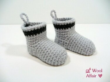 Zwei hellgraue, handgehäkelte Babysocken mit schwarzem Streifen am Schaft und kleinen Schlaufen an den Fersen vor neutralem Hintergrund.