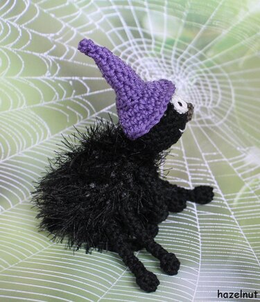 Gehäkelte schwarze Amigurumi-Spinne mit lila Hexenhut vor Spinnennetz-Hintergrund