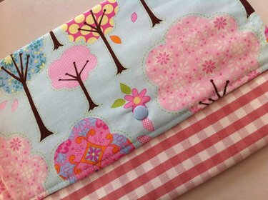 Stück Stoff mit einem Muster aus bunten Bäumen und Blumen auf hellblauem Grund, kombiniert mit rot-weiß kariertem Stoff und einem hellblauen Knopf.