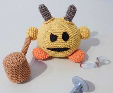 Gehäkelte runde gelbe Amigurumi-Figur mit Hörnern, orangefarbenen Füßen und gehäkeltem Hammer, Metallteile daneben