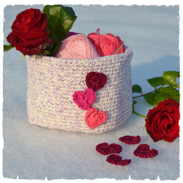 Pastellfarbener gehäkelter Utensilo mit roten Rosen, rosa Garnknäueln und Häkelherzen im Schnee