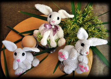 Drei weiße gehäkelte Hasen-Eierwärmer mit rosa Details auf einem Holzteller mit grünen Blättern und einem Ei