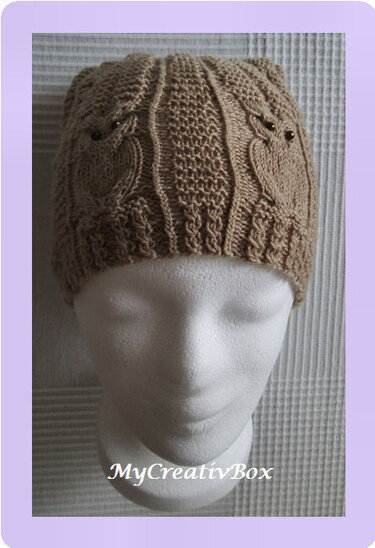 Beige gestrickte Mütze mit Eulenmotiven und Zopfmustern auf weißer Büste; Eulenaugen aus dunklen Perlen.