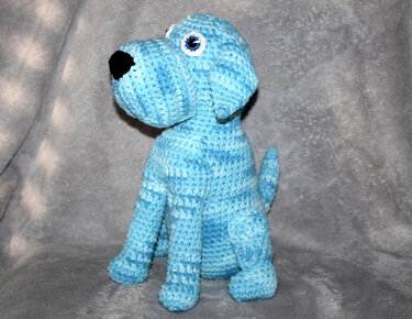 Sitzender gehäkelter Amigurumi-Hund in Blautönen mit sichtbarer Nase und Augen auf grauem Hintergrund.