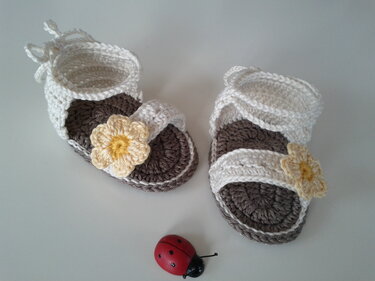 Gehäkelte Babysandalen in Creme und Braun mit gelber Blüte und Schnürband, daneben kleines Marienkäfer-Spielzeug