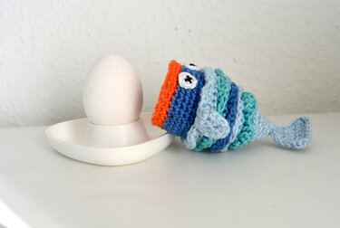 weißes Ei im Eierbecher neben gehäkeltem blauen Fisch‑Eierwärmer mit orangefarbenem Rand und Knopfaugen