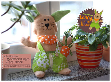 DIY-Stoffhase mit grüner Latzhose, sichtbarem Reißverschluss, Knöpfen und orange gepunktetem Halstuch auf einer Fensterbank