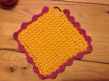 Quadratischer gelber Topflappen mit pinkfarbener Muschelkante und Aufhängeschlaufe auf Holz.