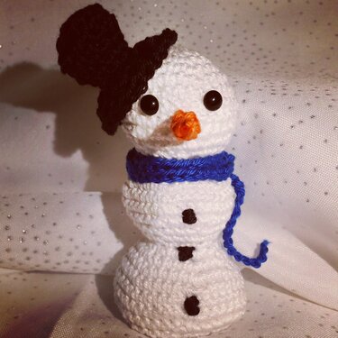 Weißer gehäkelter Amigurumi‑Schneemann mit schwarzem Hut und blauem Schal