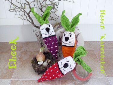 Drei gehäkelte Amigurumi‑Karotten mit Hasengesichtern in Rot, Lila und Orange mit grünen Blättern