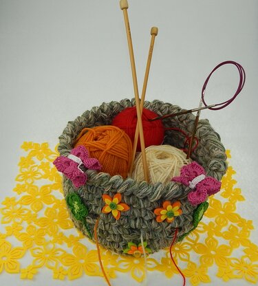 Gehäkelter runder Korb in Grau-Grün mit bunten Wollknäueln, Stricknadeln und gehäkelten Blumen- und Schmetterlingsapplikationen auf gelber Blumenunterlage