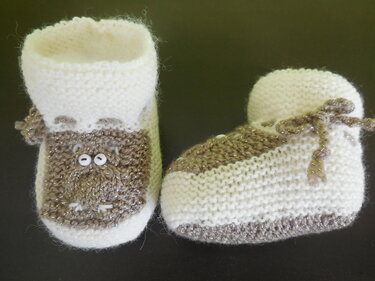 Zwei gestrickte Babyschuhe in Cremeweiß und schimmerndem Grau mit aufgesticktem Tiergesicht