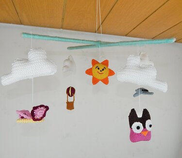 Gehäkeltes Mobile mit weißen Wolken, lächelnder Sonne, Heißluftballon, Schmetterling, Flugzeug und Eule an mintgrünen Stäben