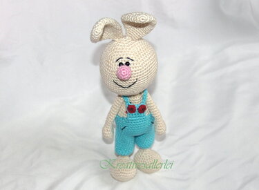 Gehäkelter hellbeiger Amigurumi-Hase mit rosa Nase und türkisblauer Latzhose auf hellem Hintergrund.