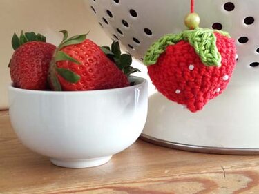 Gehäkelte Erdbeere mit grünen Blättern als Anhänger neben weißer Schale mit frischen Erdbeeren