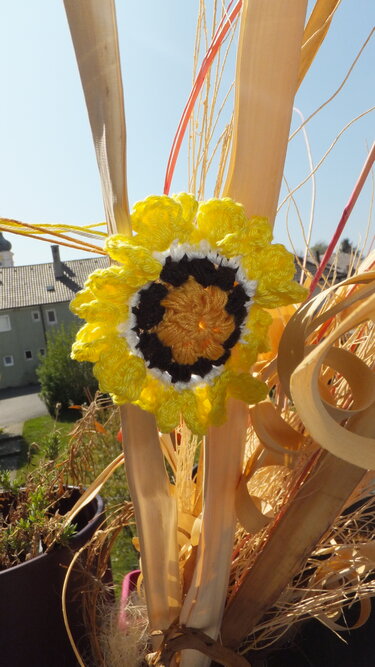 Gehäkelte gelbe Sonnenblumen-Applikation mit schwarzem Kern an getrockneten Dekorzweigen vor Häuserkulisse