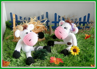 Zwei weiße und schwarze Amigurumi-Kühe mit rosa Schnauzen auf grünem Plüschgras vor blauem Miniaturzaun.