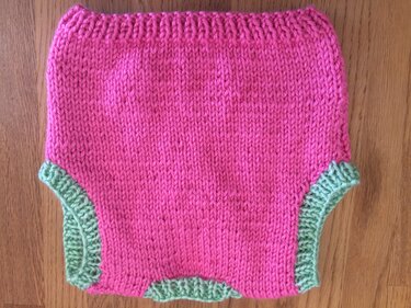 Gestrickter Baby-Pulloverrumpf in Rosa mit grünen Bündchen auf Holz