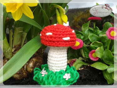 Gehäkelter roter Pilz mit weißen Punkten auf grünem Häkelboden zwischen Blumen