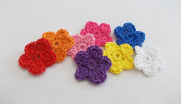 Acht gehäkelte fünfblättrige Blumen in Rot, Orange, Hellrosa, Pink, Lila, Gelb, Blau und Weiß auf hellem Hintergrund.