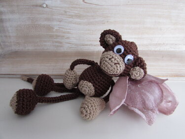 Gehäkelter brauner Amigurumi-Affe mit beigen Details, großen Kulleraugen und rosa Blütenapplikation, liegend