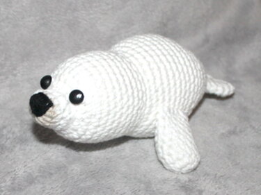 Weißes gehäkeltes Robben-Amigurumi mit schwarzen Knopfaugen und schwarzer Nase auf grauem Stoff.