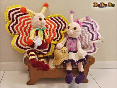 Zwei gehäkelte Amigurumi-Schmetterlinge auf einer kleinen Holzbank, einer mit gelb-orange-roten, der andere mit lila-rosa Flügeln.