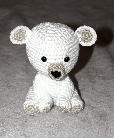 Weißes, gehäkeltes Amigurumi-Eisbärchen mit beigen Details an Ohren, Schnauze und Pfoten sowie schwarzen Augen und Nase, sitzend auf grauem Untergrund.