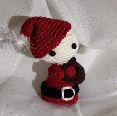 Gehäkelter Amigurumi-Weihnachtsmann mit roter Mütze, weißem Bommel, schwarzem Gürtel und braunem Sack auf glitzerndem Stoff.
