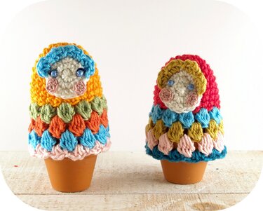 Zwei bunte handgehäkelte Eierwärmer in Puppenform auf kleinen Tontöpfen