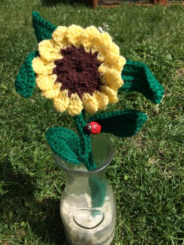 Gehäkelte gelbe Sonnenblume mit grünen Blättern und rotem Marienkäfer in einer Glasvase auf Gras