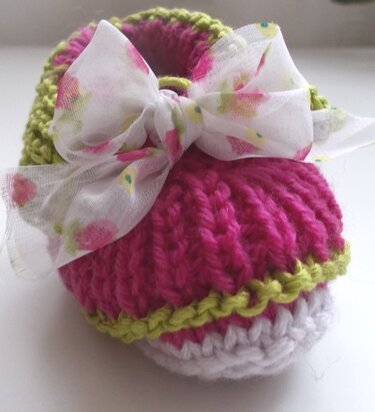 Gestrickter Babyschuh in Pink mit grünem Rand, weißer Sohle und geblümter Stoffschleife.