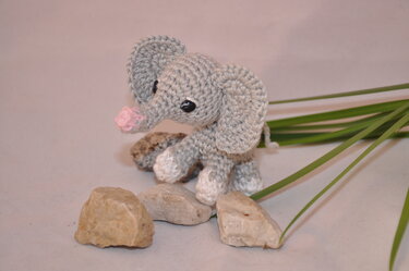 Gehäkelter Amigurumi-Elefant in Grau und Rosa, stehend zwischen Steinen und Gräsern.