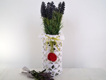 Detaillierte Ansicht einer Glasvase mit weißem, offenmaschigem Häkelüberzug im Muschelmuster und künstlichem Lavendel