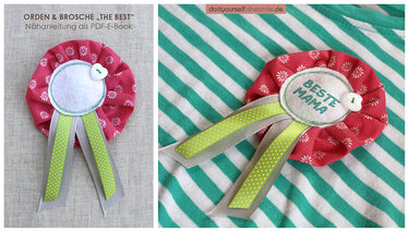 Selbstgenähte rosafarbene Orden-Brosche mit weißem Blumenmuster, grünen und grauen Bändern sowie der Aufschrift BESTE MAMA auf gestreiftem Stoff.