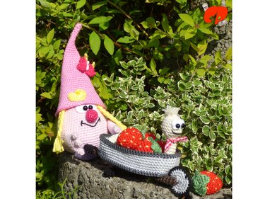 Bunter Amigurumi-Zwerg mit Wagen voller roten Erdbeeren, kleine gehäkelte Raupe mit Halstuch