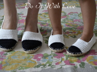 Zwei Paar gehäkelte Espadrilles in Weiß mit schwarzer Zehenkappe und Jutesohle auf floralem Teppich; ein Paar für Erwachsene und ein Paar für Kinder.