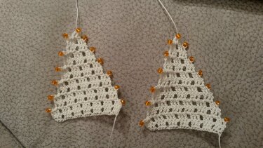 Zwei weiße, gehäkelte Weihnachtsbaumformen mit orangefarbenen Perlen an den Rändern auf grauem Untergrund