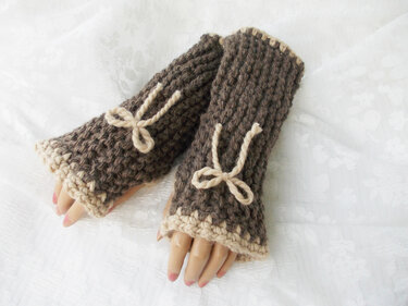 Braune, grob gestrickte fingerlose Armstulpen mit beigen Schleifen und kontrastierendem Bündchen auf hellem Hintergrund
