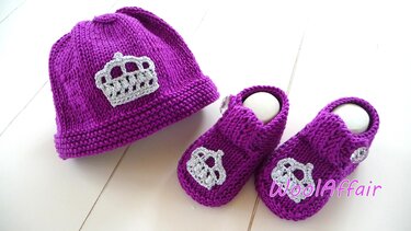 Gestrickte lilafarbene Babymütze und passende Schühchen, beide mit silberfarbener Kronenapplikation, auf hellem Untergrund.