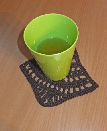 Dunkelbrauner, quadratischer Häkeluntersetzer unter einem grünen Becher auf einem Holztisch.