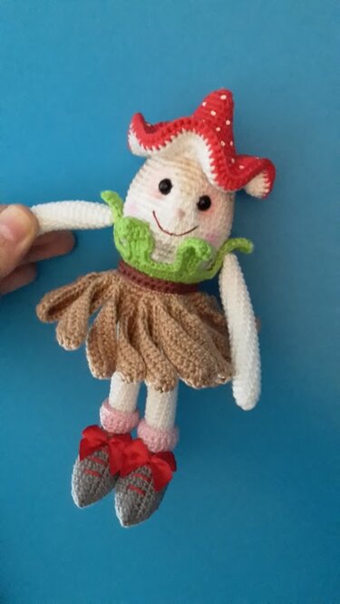 Gehäkelter Glückspilz mit rotem Tupfenhut, grünem Kragen und braunem Rock, lächelnd, von einer Hand gehalten vor blauem Hintergrund