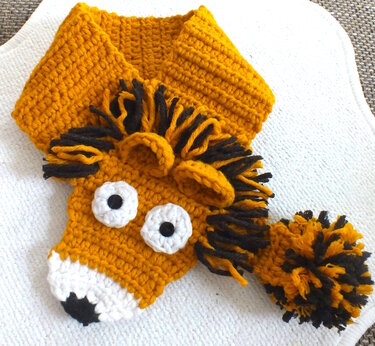 Handgehäkelter Löwenschal in Senfgelb mit schwarzer Mähne, weißen Augen und Bommel.