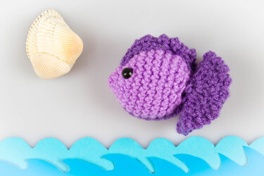 Violetter gehäkelter Amigurumi-Fisch mit schwarzem Auge neben Muschel und blauen Papierwellen