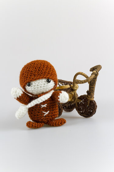 Gehäkelte Amigurumi-Puppe in Braun und Weiß neben einem kleinen Korb-Dreirad
