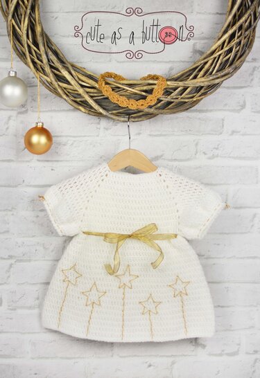 Weißes gehäkeltes Babykleid mit goldenen Sternen und goldfarbenem Satinband an einem Holzhänger vor weißer Ziegelwand.