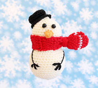 Gehäkelter Schneemann-Anhänger mit schwarzem Hut, rotem Schal und oranger Nase vor blauem Hintergrund mit Schneeflocken
